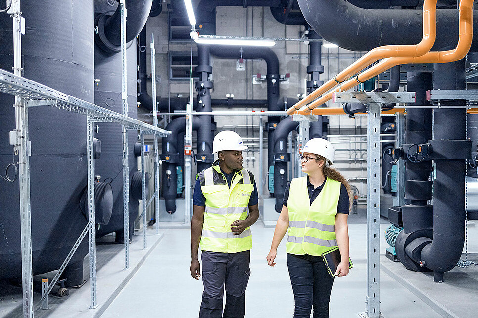 Two Leadec employee wearing safety vests and helmets walk through a technical area.