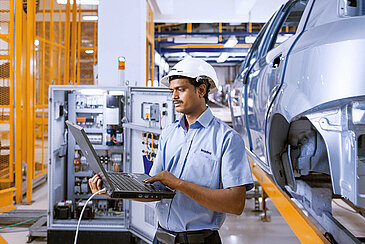 A Leadec employee with a laptop checking the power unit of a conveyor belt in an automotive factory.